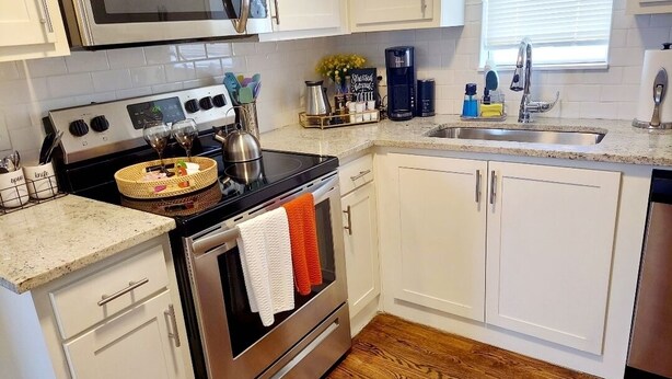 Una cocina con armarios blancos, estufa de acero inoxidable, microondas, pileta y piso de madera.
