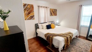 Un dormitorio con cama, mesita de noche, lámpara y una obra de arte en la pared.