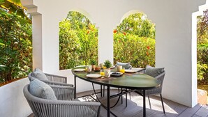 A covered outdoor dining area with a round table, wicker chairs, and a view of lush greenery through arched openings. - Genteel Home Venalmar Terrace (Estepona)
