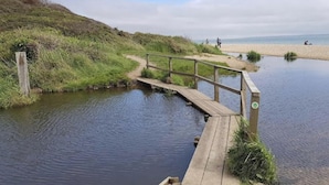 A wooden bridge over a calm body of water with a grassy bank and a sandy beach in the background. - Serene Chalet Retreat (Southampton)