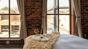 A room with a large window, a bed, a blanket, a tray with a bottle and glasses, and a view of a street.