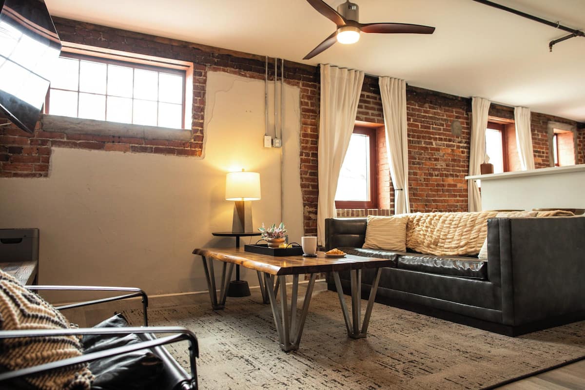 Una sala de estar con pared de ladrillo, ventilador de techo, sofá, mesa de centro y ventana con cortinas.