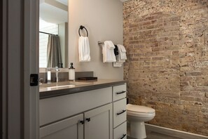 A bathroom with a brick wall, a white toilet, a sink with a mirror, and towels hanging on a rack.