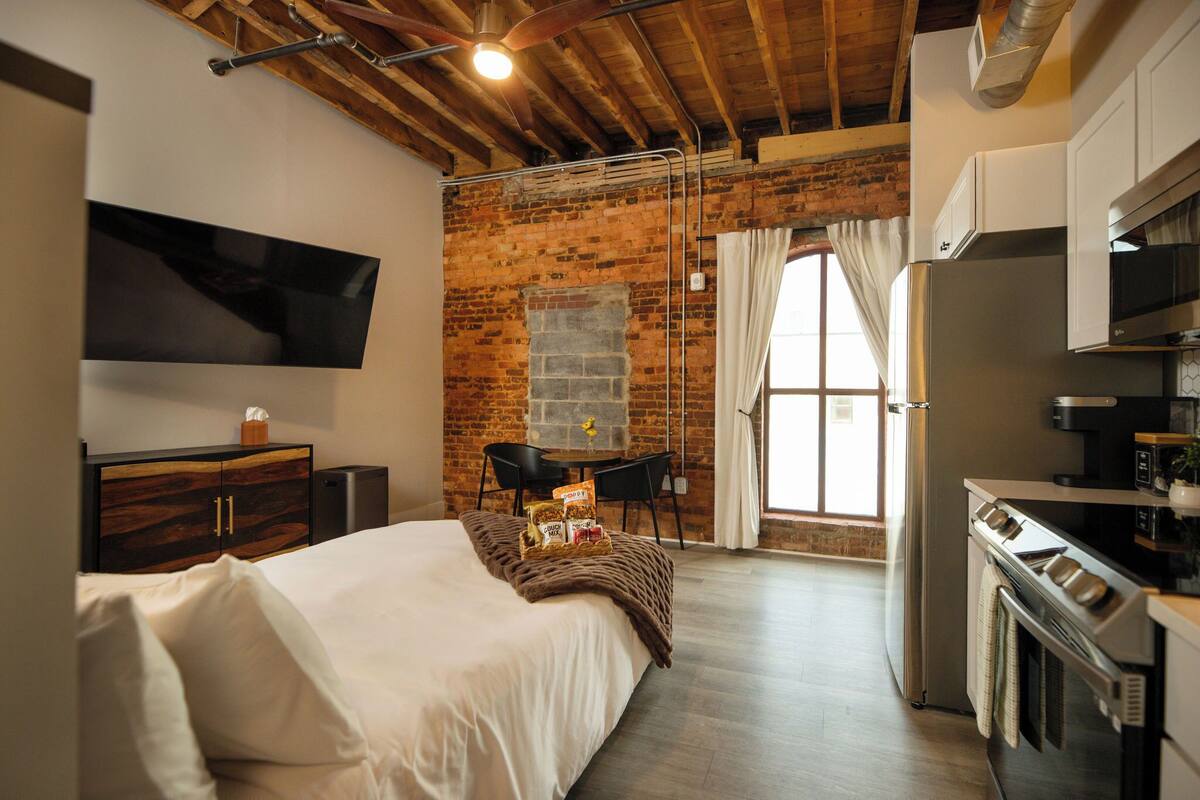 Habitación de hotel con cama, televisor montado en la pared, pared de ladrillo, ventana con cortinas y kitchenette con refrigerador y estufa.