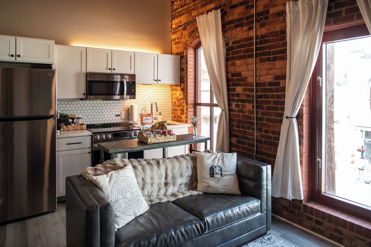 Una cocina equipada con refrigerador, microondas y estufa, una mesa de comedor con una cesta de pan, y un sofá de cuero con almohadones.