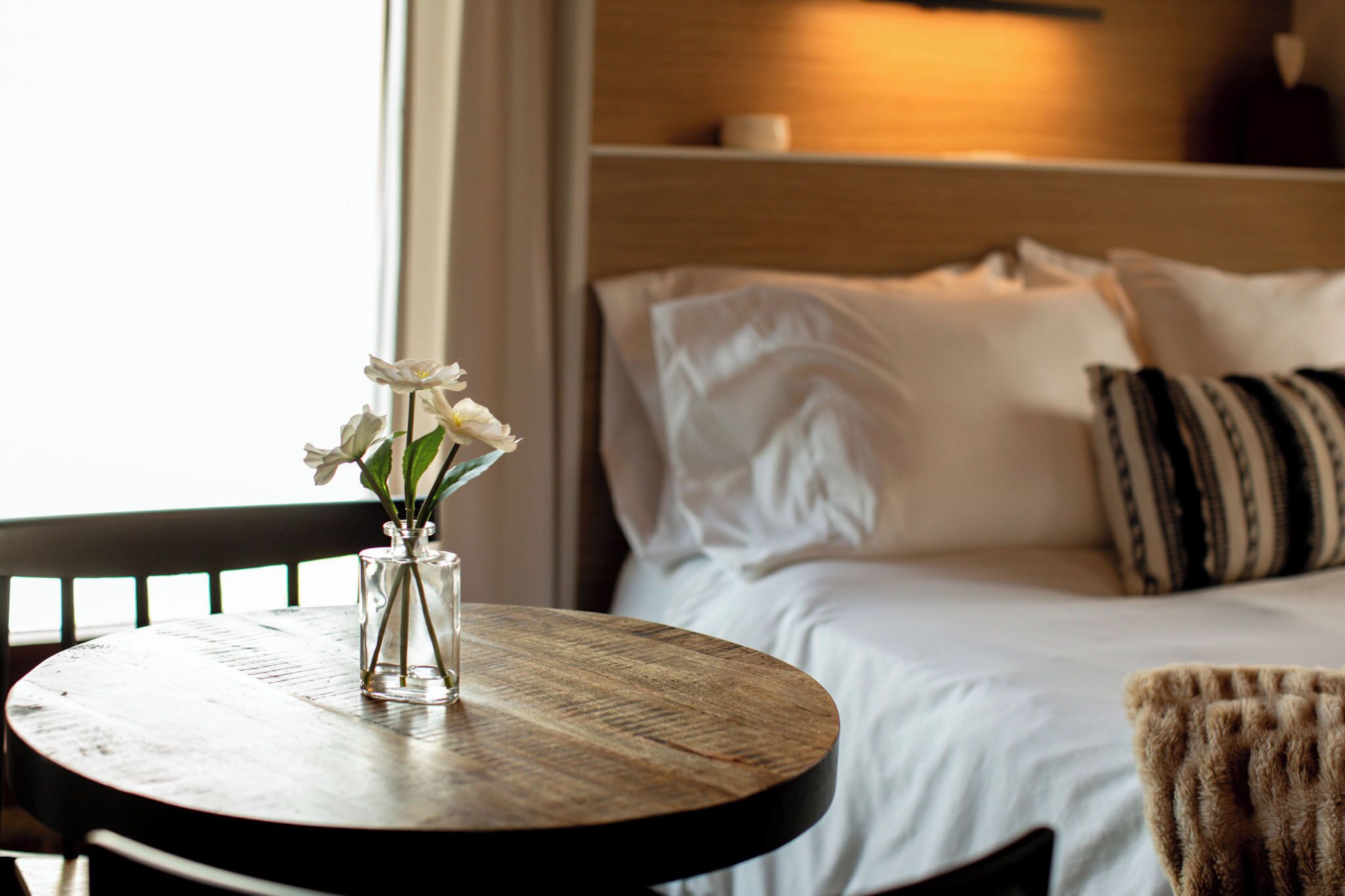 Een bed met witte lakens, een gestreept kussen en een vaas met witte bloemen op een ronde houten tafel.