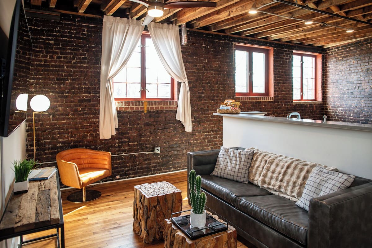 Una habitación con pared de ladrillo, techo de madera, un sofá de cuero, una mesa de centro de madera, un cactus y una pequeña mesita auxiliar.