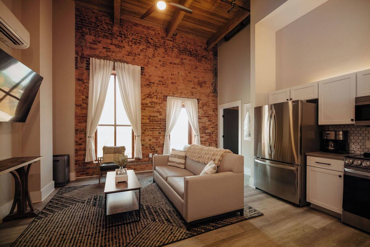 Una cocina moderna con electrodomésticos de acero inoxidable, una zona de comedor con sofá y mesa de centro, y una pared de ladrillo.