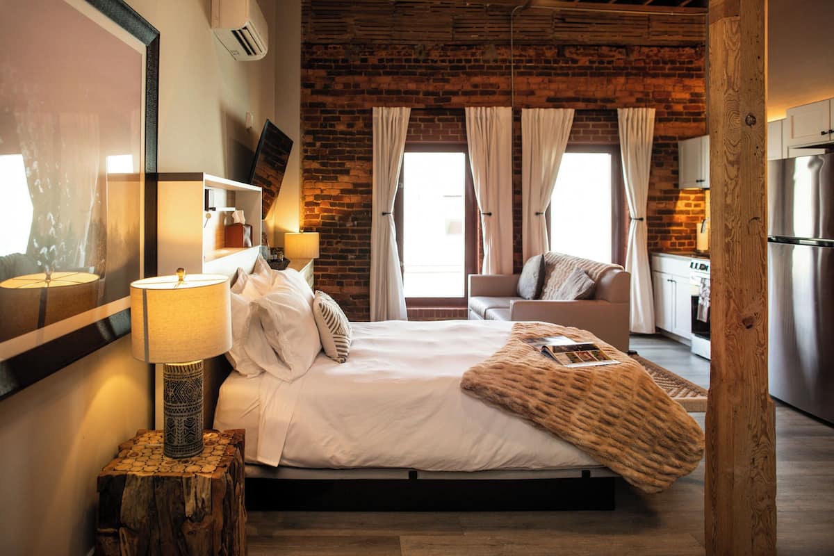 Un dormitorio con pared de ladrillo, una cama con ropa de cama blanca, una mesita de noche de madera con una lámpara y vista a la cocina.