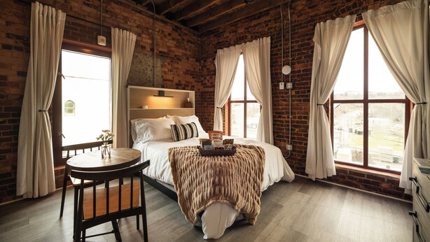 A rustic room with exposed brick walls, a wooden ceiling, a bed with a white headboard, a round wooden table, and large windows with curtains. - Three Trails Downtown Elkin (Elkin)
