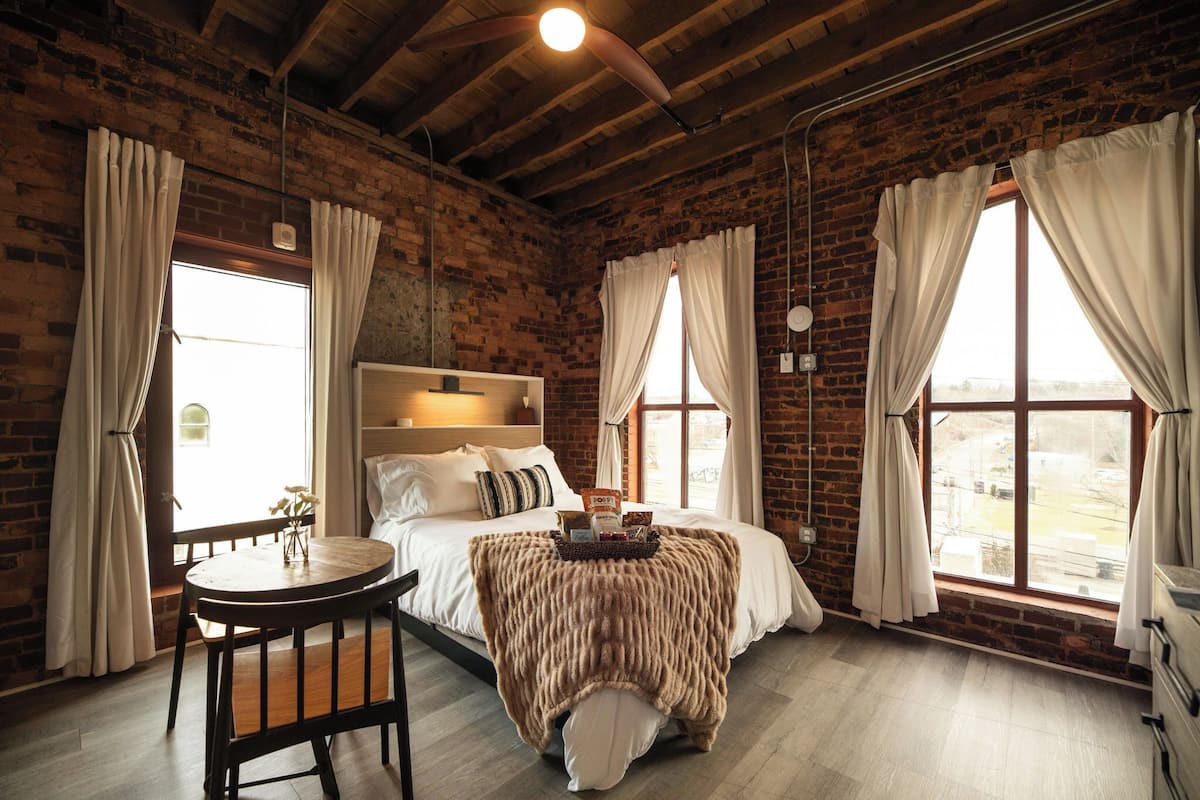 Una habitación rústica con paredes de ladrillo visto, techo de madera, una cama con cabecera blanca, una mesa redonda de madera y ventanas grandes con cortinas.