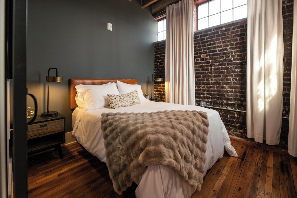 Un dormitorio con pared de ladrillo, suelo de madera, cama con ropa blanca, mesita de noche con lámpara y ventana con cortinas.
