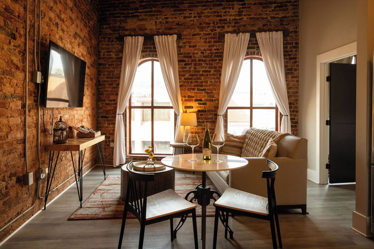 Una habitación acogedora con pared de ladrillo, una mesa redonda con sillas, un sofá y un televisor montado en la pared.