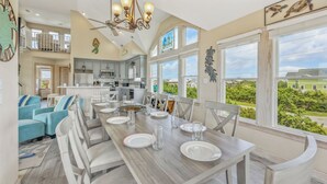 A spacious dining area with a long table set for six, surrounded by white chairs. A kitchen with modern appliances is visible in the background, and large windows offer a view of the greenery outside. - Soundside Dog Friendly Home w/ Pool, Hot Tub, Game Room, Tiki Bar, Public Dock (Rodanthe)