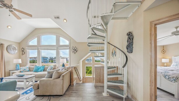 A spacious living area with a high ceiling, a spiral staircase, and large windows offering a view of the outside. - Soundside Dog Friendly Home w/ Pool, Hot Tub, Game Room, Tiki Bar, Public Dock (Rodanthe)