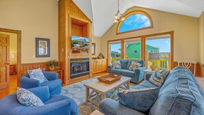 A spacious living room with a fireplace, blue sofas, and a large window overlooking a green house. - Soundside Dog Friendly Home w/ Private Pool, HotTub, Game Room, Community Tennis (Avon)