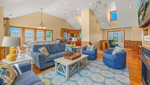 A spacious living area with a large sofa, a smaller blue armchair, and a coffee table. There is a kitchen area in the background with wooden cabinets and a dining table. - Soundside Dog Friendly Home w/ Private Pool, HotTub, Game Room, Community Tennis (Avon)