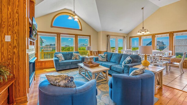 A spacious living room with a high ceiling, large windows, and a cozy seating area with blue sofas and a coffee table. - Soundside Dog Friendly Home w/ Private Pool, HotTub, Game Room, Community Tennis (Avon)