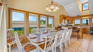 A spacious dining area with a large table and white chairs, a kitchen with wooden cabinets, and large windows offering a view of the outside. - Soundside Dog Friendly Home w/ Private Pool, HotTub, Game Room, Community Tennis (Avon)