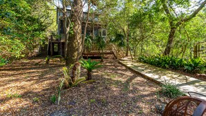 Una casa en los árboles con un porche de madera y escaleras que llevan a ella, rodeada de vegetación densa y un sendero de hormigón.