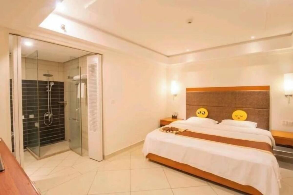 A hotel room with a bed, a bedside table, a nightstand, a glass-enclosed shower, and a door leading to another room.
