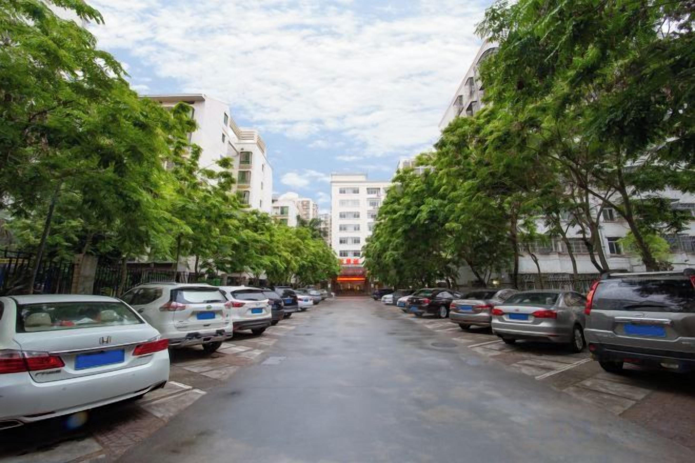 Una calle bordeada por coches aparcados y árboles, que conduce a un edificio con un letrero rojo.