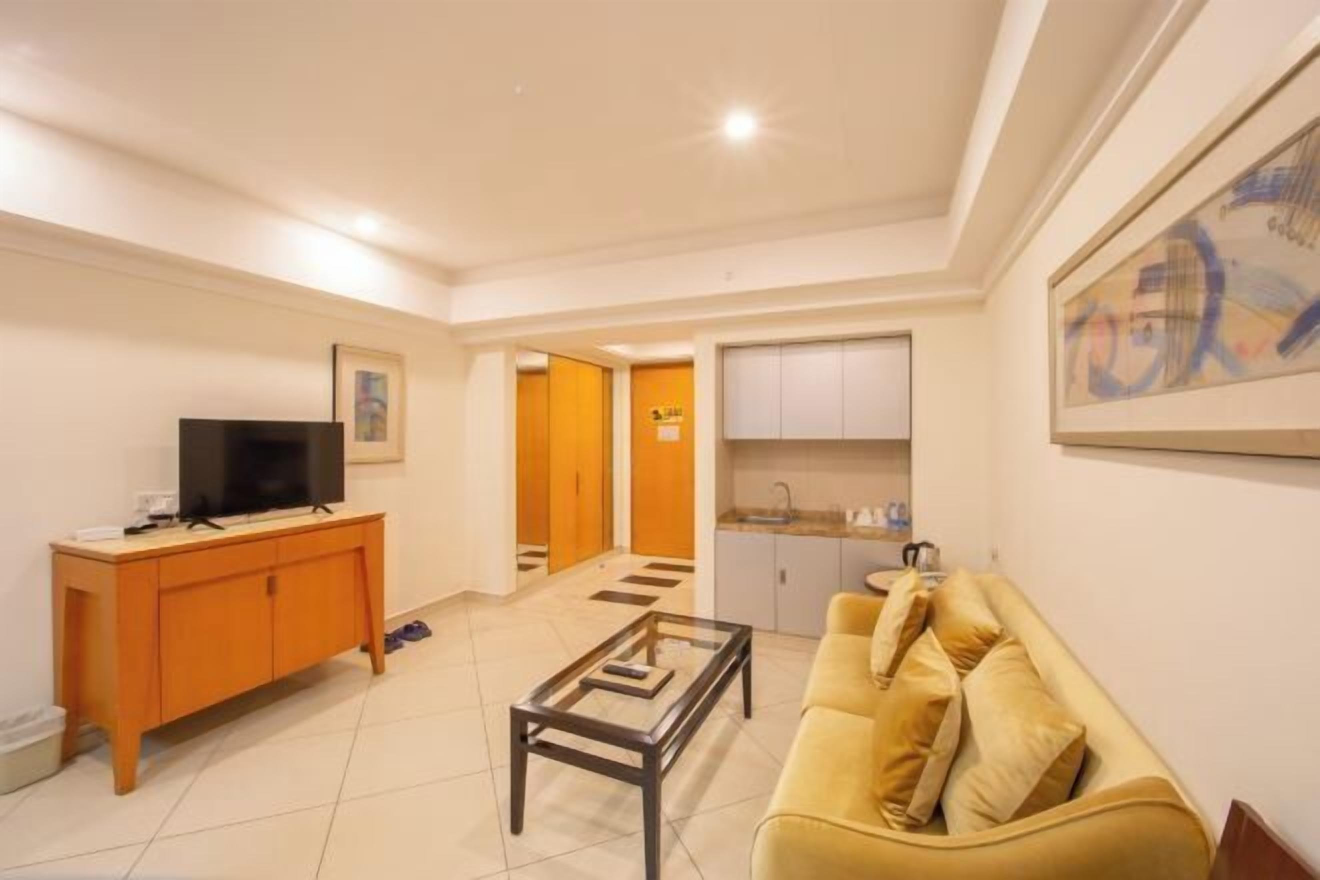 A living room with a sofa, a coffee table, a TV, and a cabinet. There is a kitchen area with cabinets and a sink.