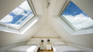 Een kleine, driehoekige kamer met twee bedden, een tafeltje en twee lampen. Er zijn dakramen in het plafond, waardoor natuurlijk licht binnenkomt.