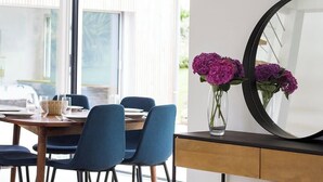 Een eethoek met een tafel voor vier personen, blauwe stoelen, een vaas met paarse bloemen en een grote spiegel aan de muur.