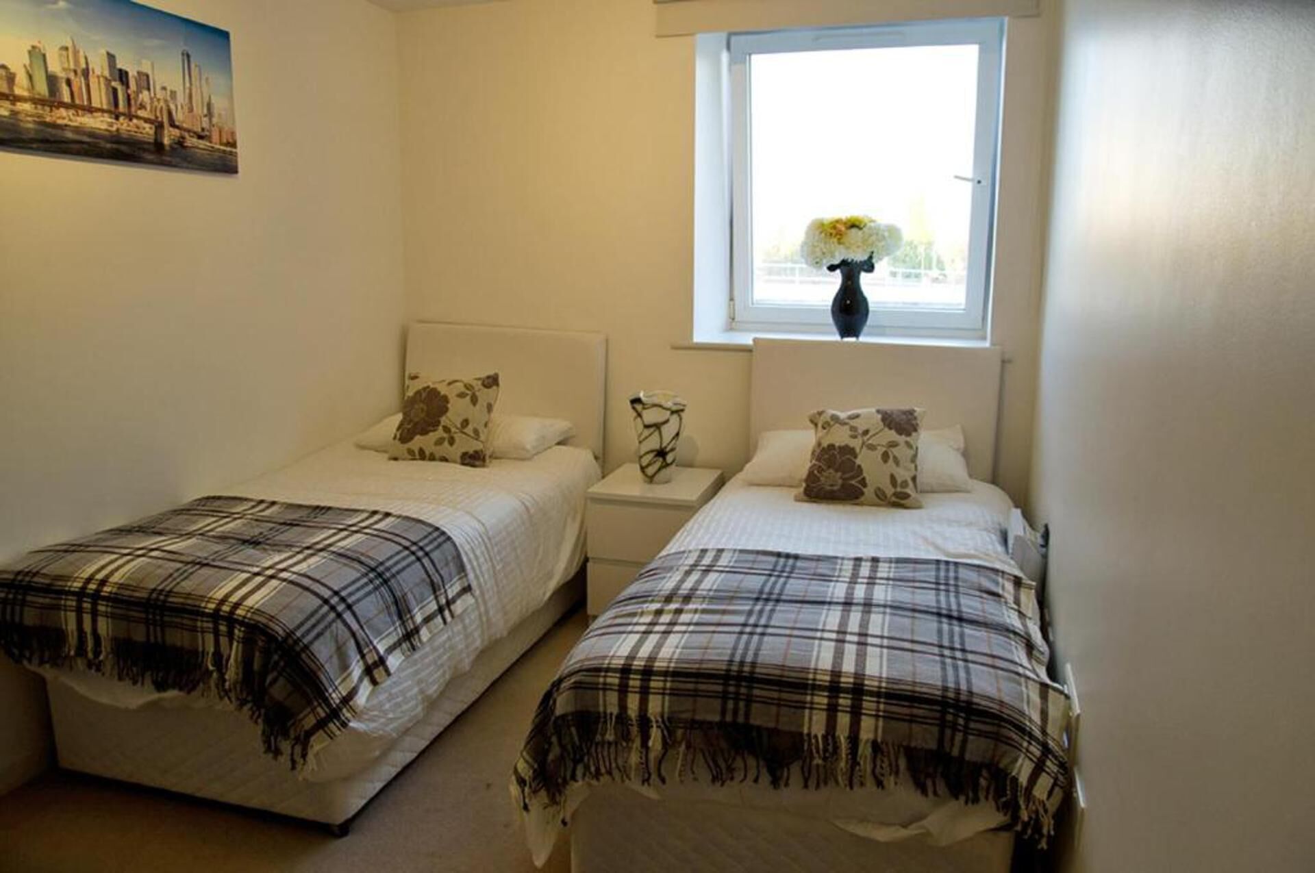 A bedroom with two beds, each with a plaid bedspread and matching pillows. There is a window with a view of a cityscape and a vase with flowers on the windowsill.