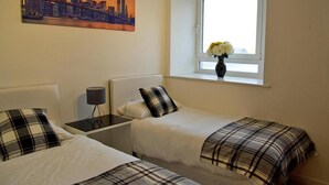 A hotel room with two beds, a nightstand, a lamp, a window with flowers, and a cityscape painting on the wall.