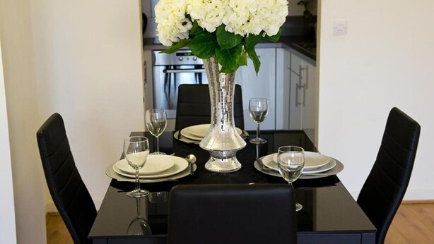 A dining table set with white plates, silverware, and wine glasses, centered with a vase of white flowers.
