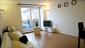 A modern living room with a sofa, a TV on a white stand, and a large window with a view of buildings.