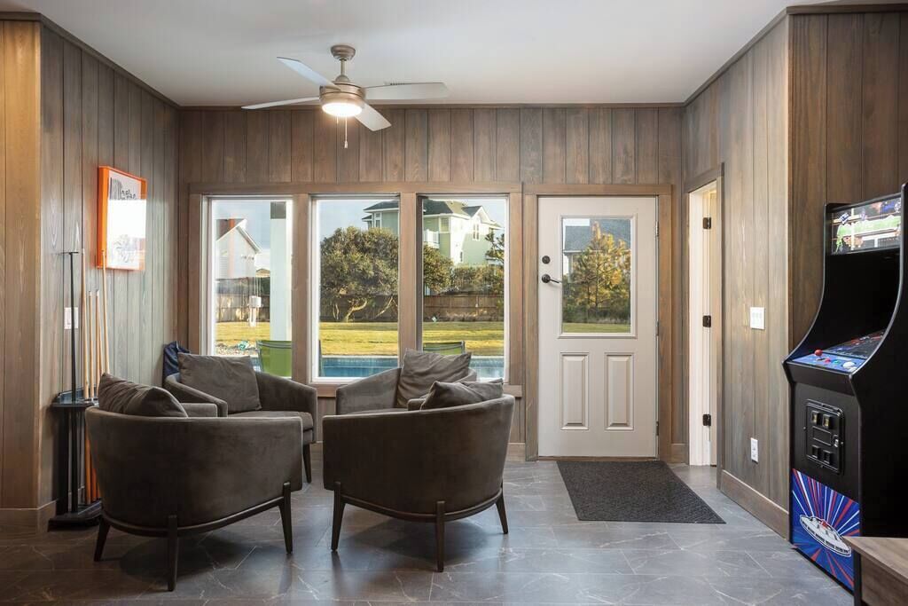Ein Zimmer mit zwei dunkelbraunen Sesseln, einem Deckenventilator, einem großen Fenster mit Blick auf einen Pool und Häuser sowie einem Spielgerät.