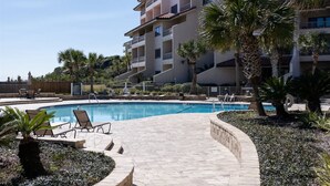 Une piscine entourée de chaises longues, de palmiers et, au fond, un immeuble à plusieurs étages.