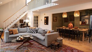 En rummelig stue med en grå sofa, et sofabord og et spiseområde med et træbord og stole. En trappe med hvide gelændere fører op til en etage.