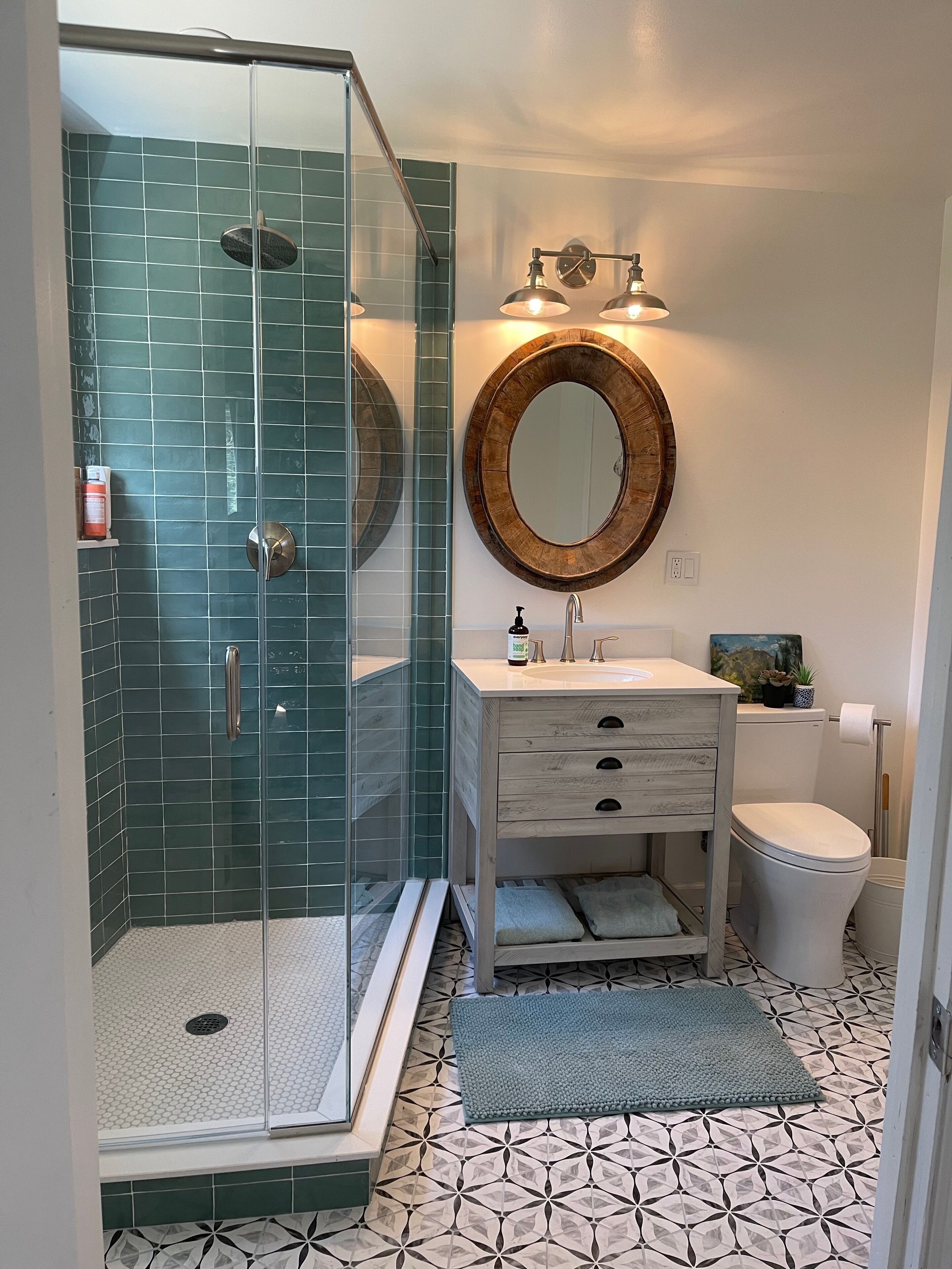 A bathroom with a glass-enclosed shower, a round mirror, a sink with a wooden frame, and a toilet.