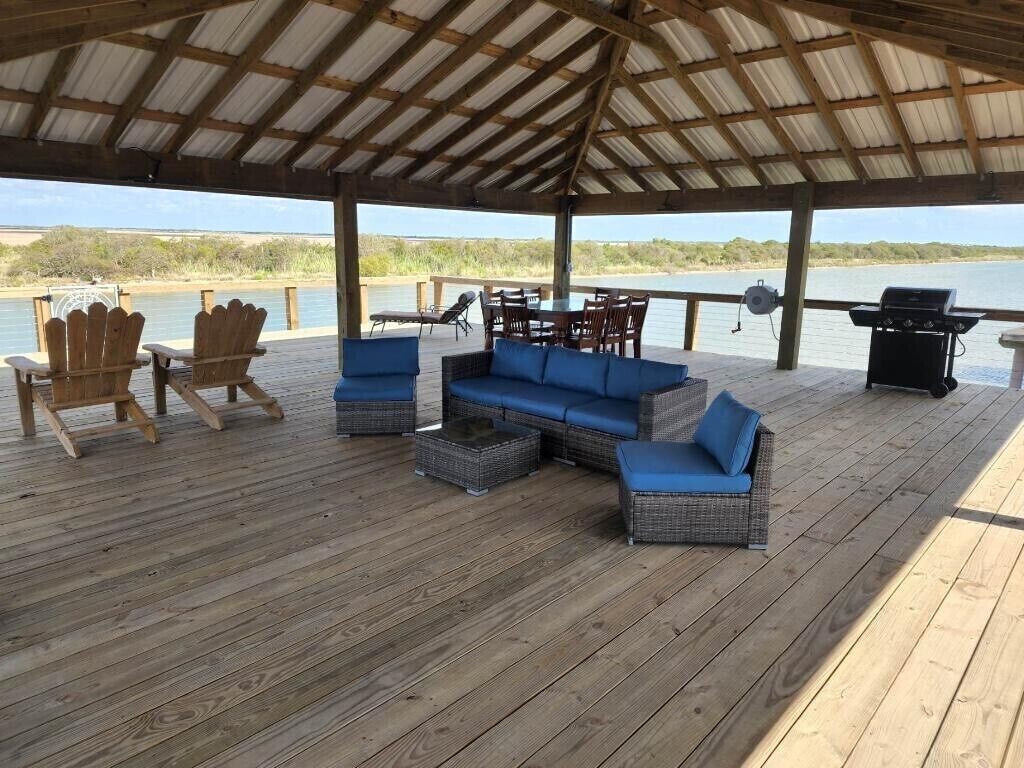 Une terrasse extérieure couverte, aménagée avec des meubles en osier, un barbecue et offrant une vue sur un plan d’eau.