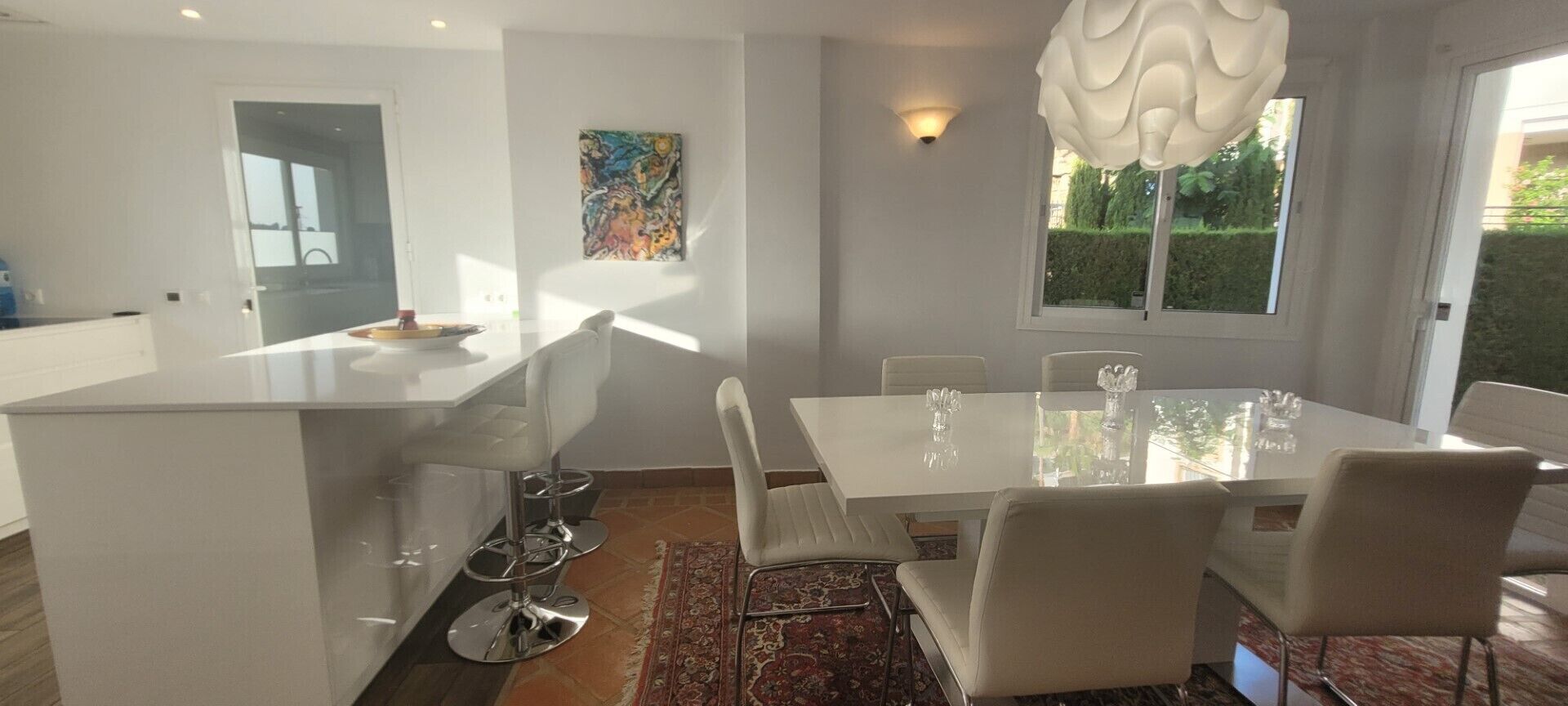 Et moderne køkken med en central ø, hvide barstole og et spiseområde med et hvidt bord og stole.