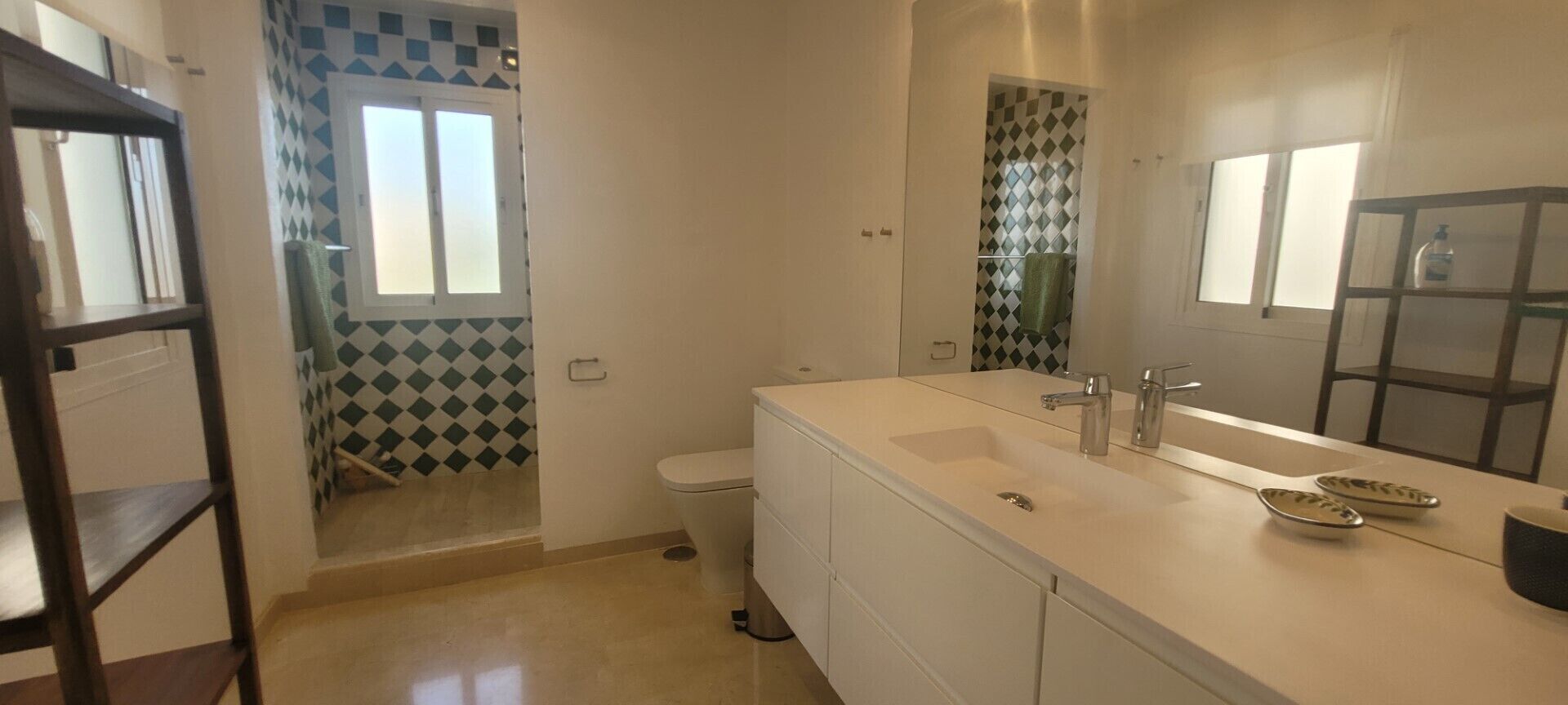 Un baño con lavamanos blanco, espejo y retrete. Se aprecia una ventana con diseño de cuadros y una puerta con un diseño similar.