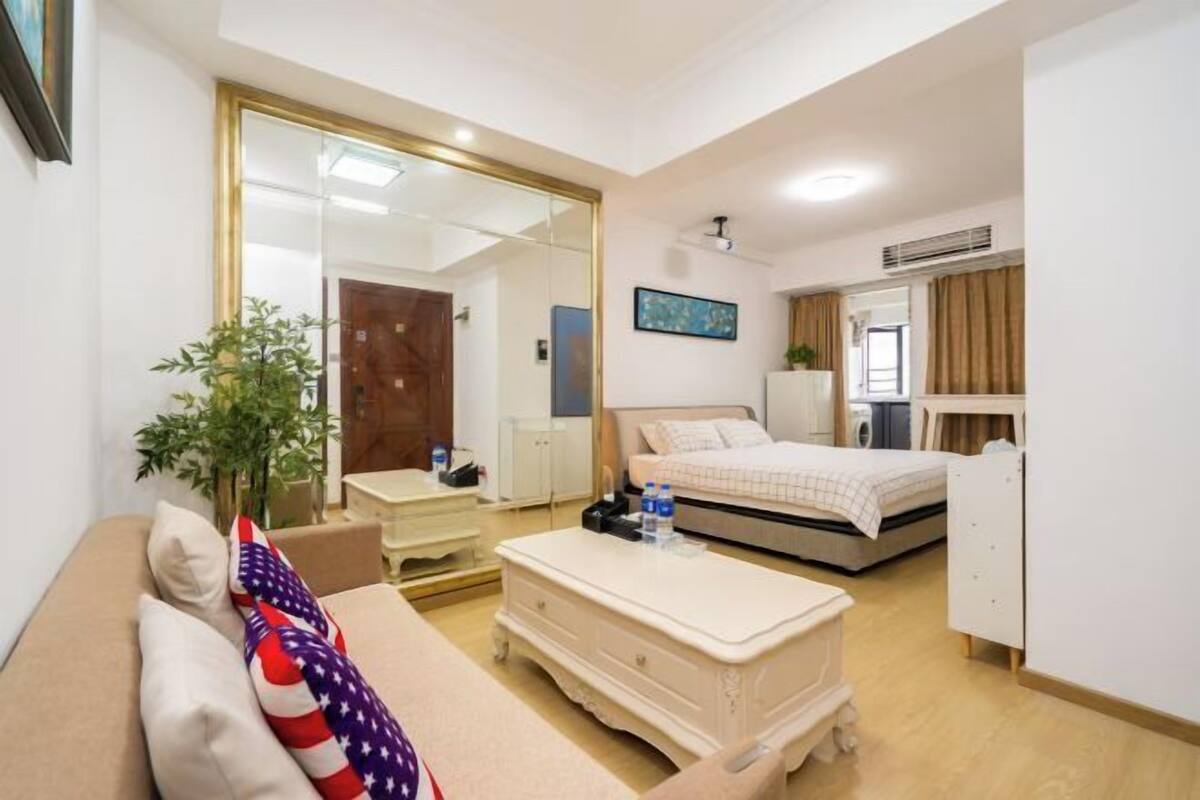 Une chambre d’hôtel avec un lit, une table de chevet avec des bouteilles d’eau, un canapé avec un coussin orné d’un drapeau, un miroir, une armoire et une petite plante.