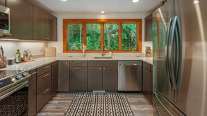 A modern kitchen with stainless steel appliances, a large window with a view of greenery, and a patterned rug.