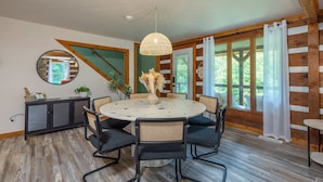 A dining area with a round table, black chairs, a woven pendant light, and a view of the outdoors through large windows.