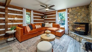 A cozy cabin interior with a wooden log wall, a sectional sofa, a round coffee table, and a fireplace.