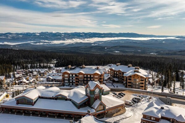 A snow-covered resort with multiple buildings, including a lodge with a clock tower, surrounded by mountains and pine forests.