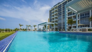 A swimming pool with a blue tiled edge, surrounded by a modern building with balconies and a covered seating area. - Ground floor Oceanfront Studio to Relax and Unwind in Aruba! (Noord)