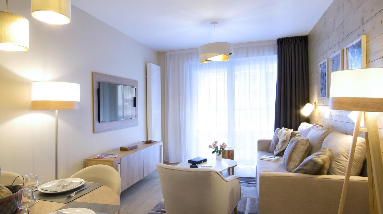 A modern hotel room with a sofa, dining table, and a TV mounted on the wall.