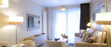 A modern hotel room with a sofa, dining table, and a TV mounted on the wall.