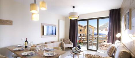 A modern hotel room with a dining area, a sofa set, and a large window offering a view of the city.