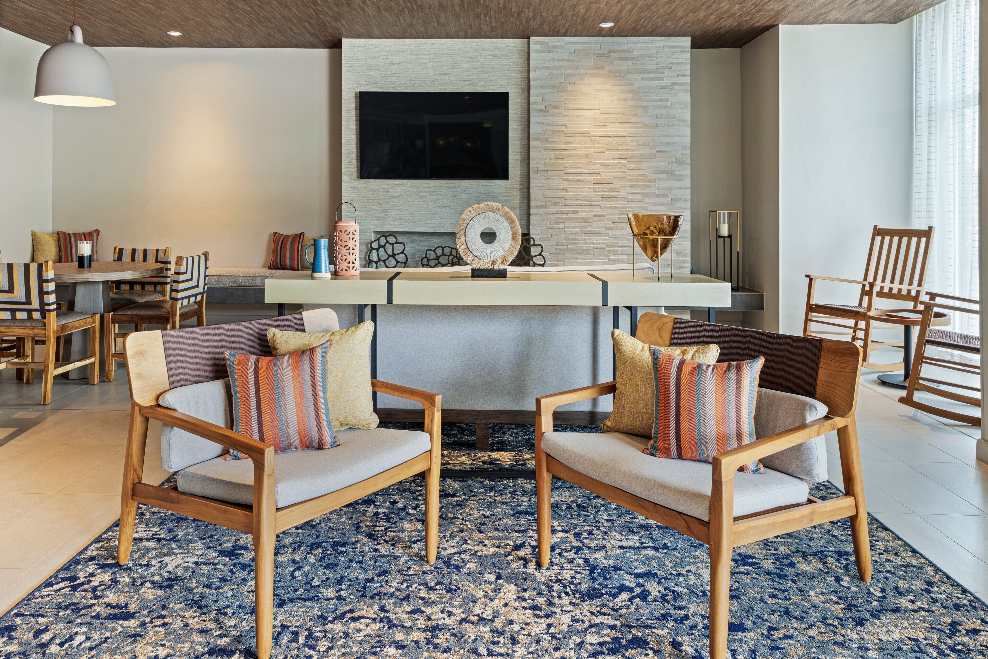 A modern living room with two wooden chairs, a blue patterned rug, and a flat-screen TV mounted on the wall.